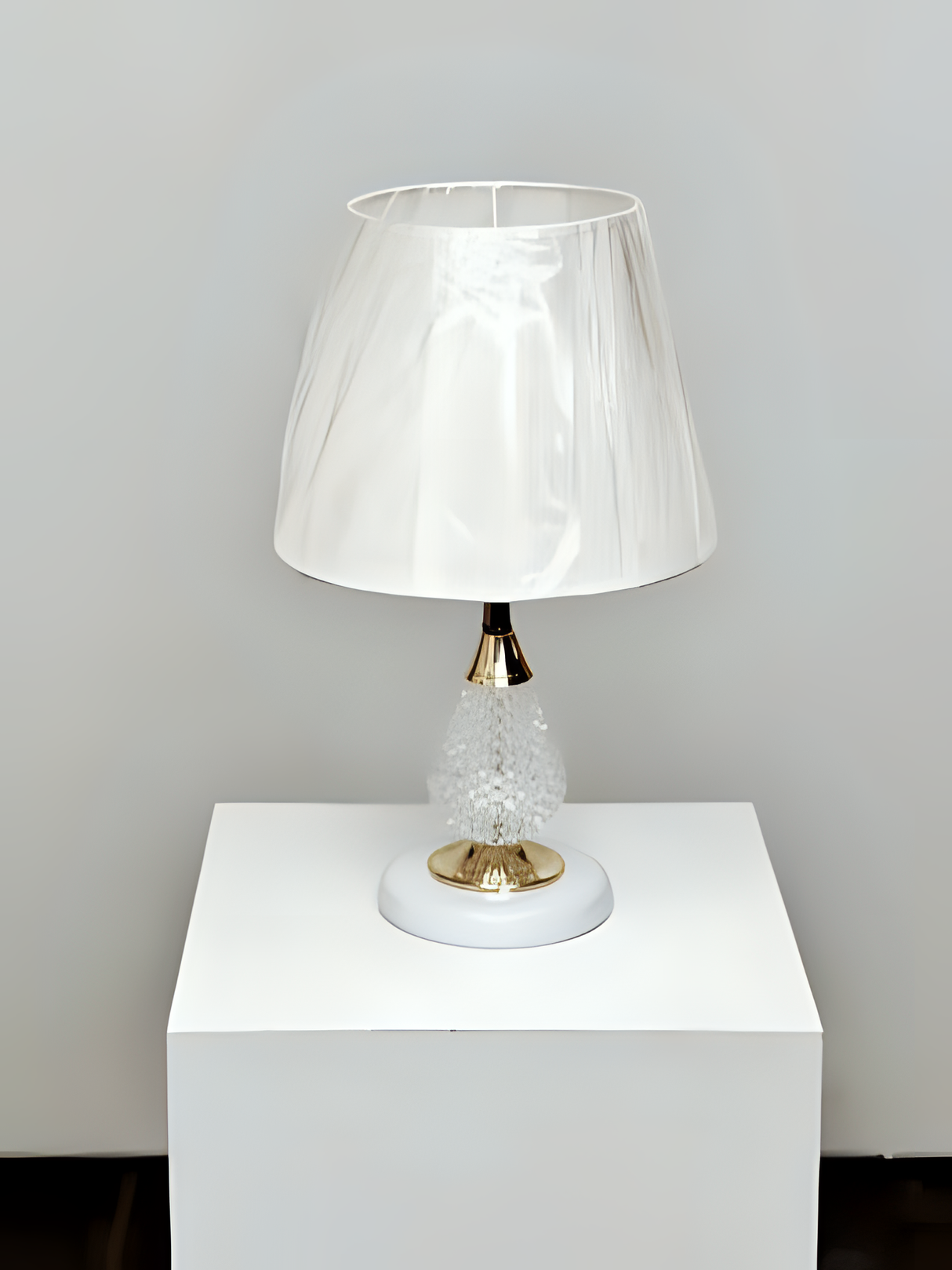 A table lamp with a crystal-textured white metal base, gold accents, and a soft beige fabric lampshade, placed on a white table against a white wall.
