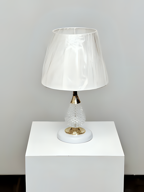 A table lamp with a crystal-textured white metal base, gold accents, and a soft beige fabric lampshade, placed on a white table against a white wall.