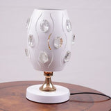A white globe table lamp with a geometric design, adorned with crystal beads, gold accents, and a sleek base, sits on a wooden table. It features an LED light with an E27 bulb, casting a warm, stylish glow.
