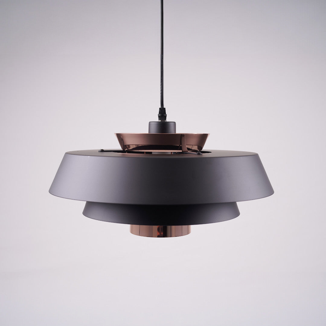 A modern layered pendant light with a matte black and copper finish, featuring a wide circular design with concentric discs, hanging from a black cord against a light gray background.