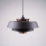 A modern layered pendant light with a matte black and copper finish, featuring a wide circular design with concentric discs, hanging from a black cord against a light gray background.