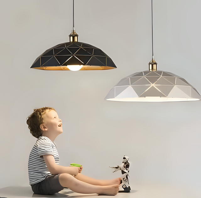 Two geometric pendant lights with black and white shades, featuring gold accents, hanging above a young boy holding a toy and looking up at the lights, with a small robot toy beside him.