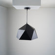 A geometric black pendant light with a faceted, polygonal design is suspended from a white ceiling. The fixture is fixed directly to a square ceiling mount.
