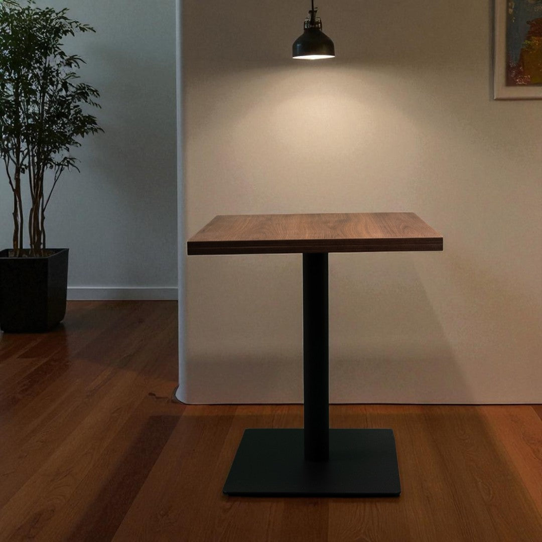Wooden table with black base in a room with a plant and light fixture.