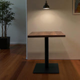 Wooden table with black base in a room with a plant and light fixture.