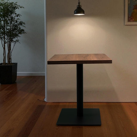 Wooden table with black base in a room with a plant and light fixture.