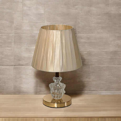 Classic gold and glass table lamp with a pleated beige lampshade and an ornate crystal base.

