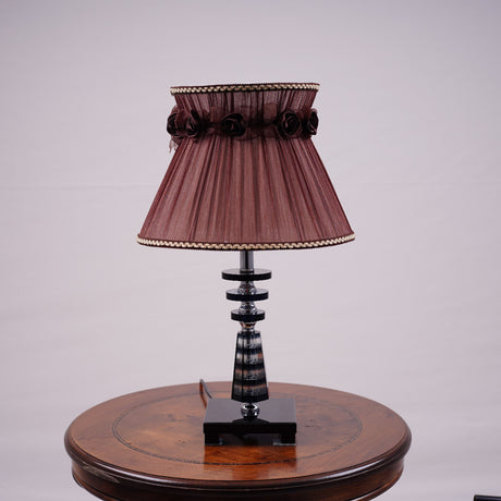 Elegant table lamp with a Brown pleated lampshade, adorned with delicate fabric roses and gold trim. The base is crafted from a combination of transparent and amber-colored glass with a geometric design, supported by a sturdy glass platform. Equipped with an E27 LED light, it emits a warm and sophisticated glow. Placed on a round wooden table.