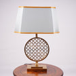 A modern table lamp featuring a gold metal geometric base with intricate latticework, placed on a round wooden table. It is complemented by a rectangular aqua-colored fabric shade with delicate gold trim. Equipped with an E27 LED light, it emits a warm and sophisticated glow.