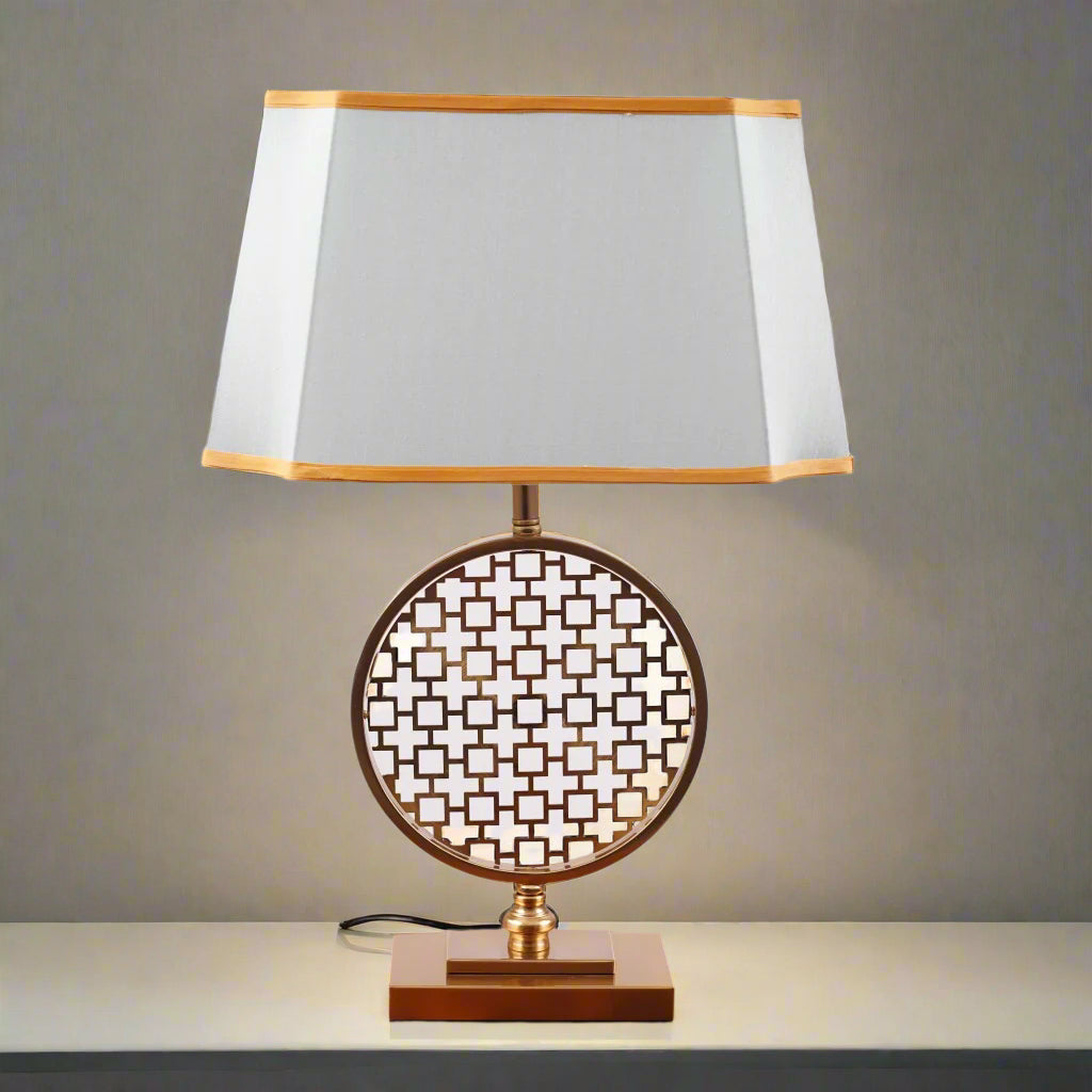 A modern table lamp featuring a gold metal geometric base with intricate latticework, placed on a round wooden table. It is complemented by a rectangular aqua-colored fabric shade with delicate gold trim. Equipped with an E27 LED light, it emits a warm and sophisticated glow.