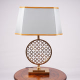 A modern table lamp featuring a gold metal geometric base with intricate latticework, placed on a round wooden table. It is complemented by a rectangular aqua-colored fabric shade with delicate gold trim. Equipped with an E27 LED light, it emits a warm and sophisticated glow.