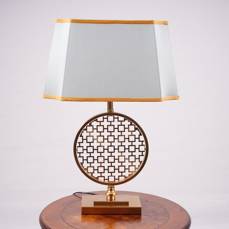 A modern table lamp featuring a gold metal geometric base with intricate latticework, placed on a round wooden table. It is complemented by a rectangular aqua-colored fabric shade with delicate gold trim. Equipped with an E27 LED light, it emits a warm and sophisticated glow.
