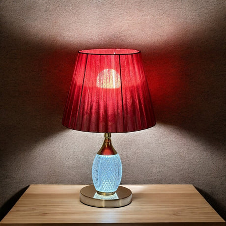 Bold red and gold table lamp with a striking red pleated lampshade equipped with an E27 LED bulb. The base features a gold finish with a built-in LED light inside the glass, adding a warm glow, placed on a wooden table.