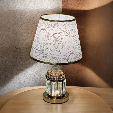 A vintage-style table lamp with a gold and glass base, and a white lampshade featuring a modern circular pattern, placed on a wooden surface with soft lighting.