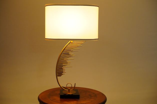 A contemporary table lamp featuring a gracefully curved gold metal base with cascading gold rods, mounted on a glossy black square platform. It is topped with a cylindrical white fabric shade with brown trim. Equipped with an E27 LED bulb, it emits a warm, ambient glow, enhancing its elegant and modern design