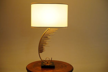 A contemporary table lamp featuring a gracefully curved gold metal base with cascading gold rods, mounted on a glossy black square platform. It is topped with a cylindrical white fabric shade with brown trim. Equipped with an E27 LED bulb, it emits a warm, ambient glow, enhancing its elegant and modern design