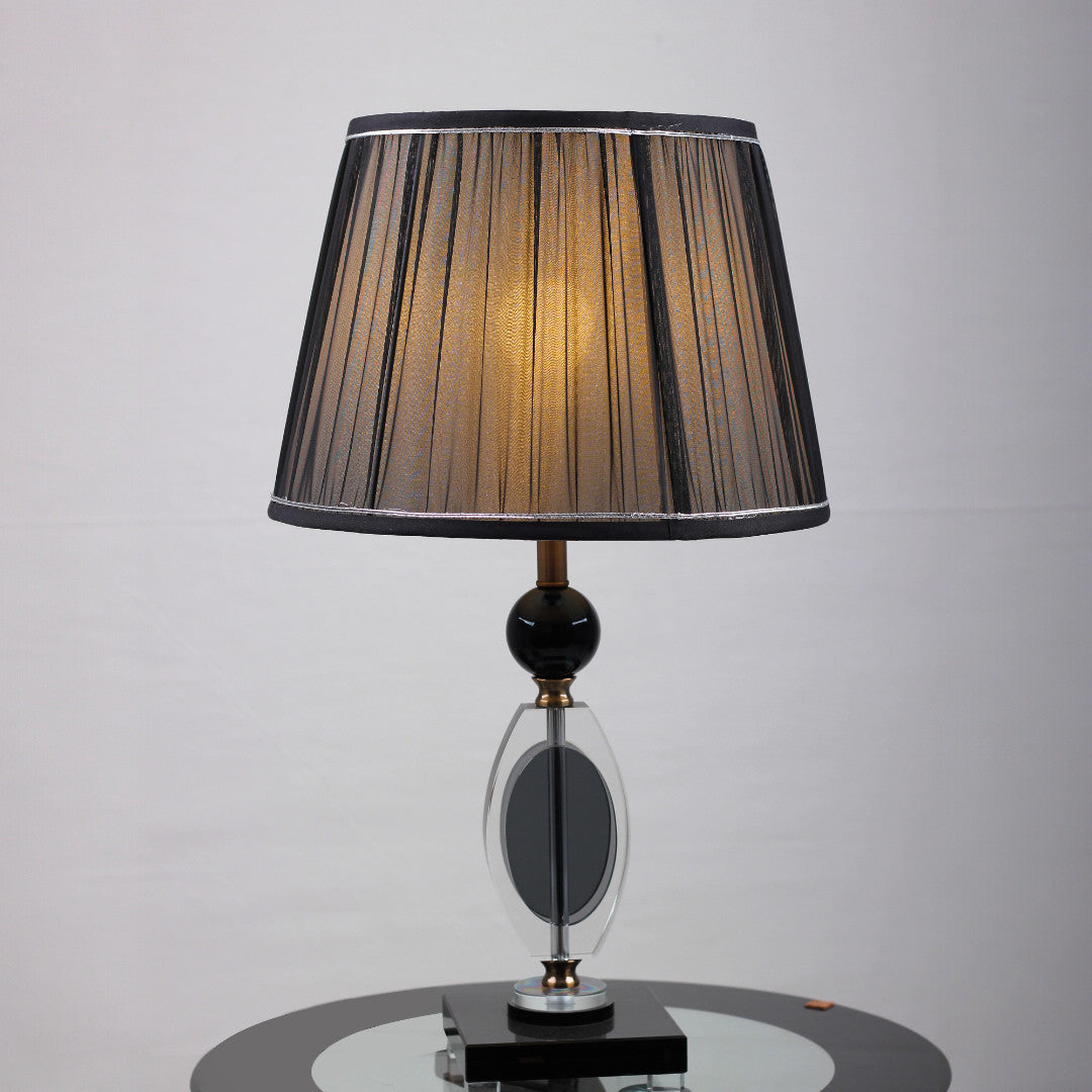 An elegant table lamp with a black pleated lampshade, equipped with an E27 LED light, emits a warm and sophisticated glow. The base is crafted from a combination of gold metal and transparent black glass with a geometric design, supported by a sturdy black glass platform.  placed on a round glass table.