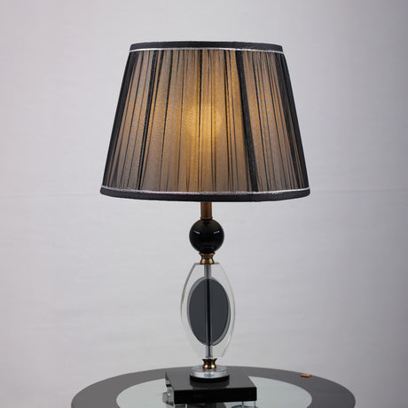 An elegant table lamp with a black pleated lampshade, equipped with an E27 LED light, emits a warm and sophisticated glow. The base is crafted from a combination of gold metal and transparent black glass with a geometric design, supported by a sturdy black glass platform.  placed on a round glass table.