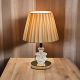 Warm-toned table lamp with an beige color pleated shade and a glass and gold base, placed on a wooden table.