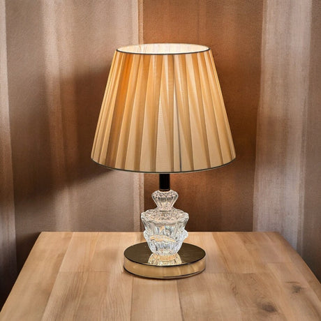 Warm-toned table lamp with an beige color pleated shade and a glass and gold base, placed on a wooden table.