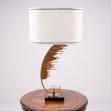 A contemporary table lamp with a gold metal base shaped like a curved staircase or an abstract wave pattern. The base is mounted on a black square platform. The lamp has a large, cylindrical white fabric shade with a thin brown trim at the edges. It is placed on a round wooden table, creating a stylish contrast.
