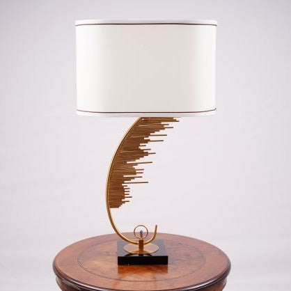 A contemporary table lamp with a gold metal base shaped like a curved staircase or an abstract wave pattern. The base is mounted on a black square platform. The lamp has a large, cylindrical white fabric shade with a thin brown trim at the edges. It is placed on a round wooden table, creating a stylish contrast.