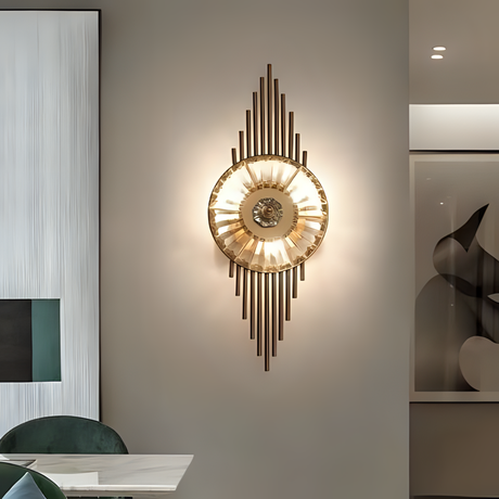 A luxurious gold and crystal wall sconce with vertical metal rods extending above and below a central circular light fixture, emitting a soft glow. mounted in a modern interior with a minimalist design, positioned near a dining area.