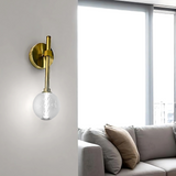 A modern wall sconce with a gold metal base and glass globe shade mounted on a white wall. In the background, a bright living room features a beige sofa with multiple throw pillows, sheer white curtains, and a white coffee table with stacked magazines.