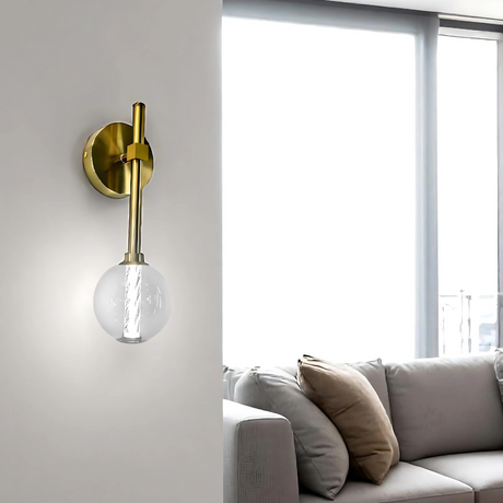 A modern wall sconce with a gold metal base and glass globe shade mounted on a white wall. In the background, a bright living room features a beige sofa with multiple throw pillows, sheer white curtains, and a white coffee table with stacked magazines.