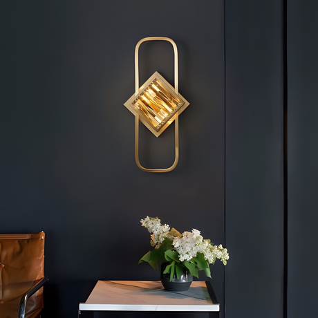 A luxurious gold wall sconce with an elongated oval frame and a central diamond-shaped glass accent, mounted on a dark wall near a side table.