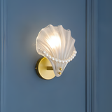 A wall-mounted seashell-shaped light fixture with a gold circular base, installed on a dark blue wall, featuring a white glass shell with pearl-like details.